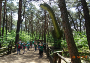 Dzieci idą ścieżką w Parku Dinozaurów. Po prawej stronie postać dinozaura.